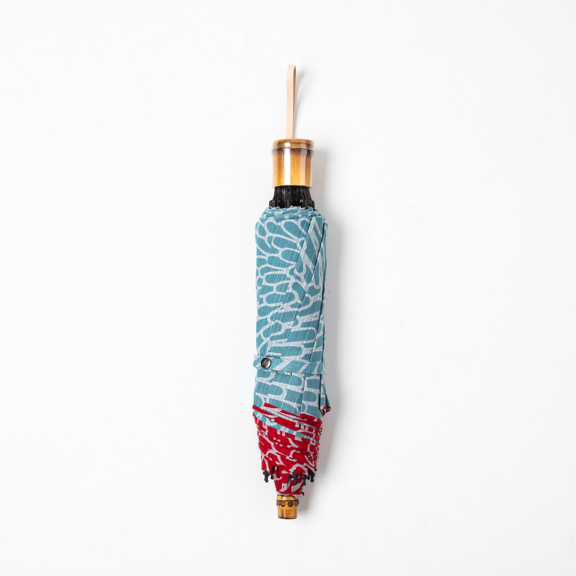 Isa Moyō Folding Umbrella in its compact, folded state, highlighting the bamboo handle and the contrast between the outer red fabric and inner light blue lining.