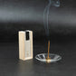 Sandalwood incense stick burning in a glass holder beside a light wooden box with black and beige label on a dark stone background