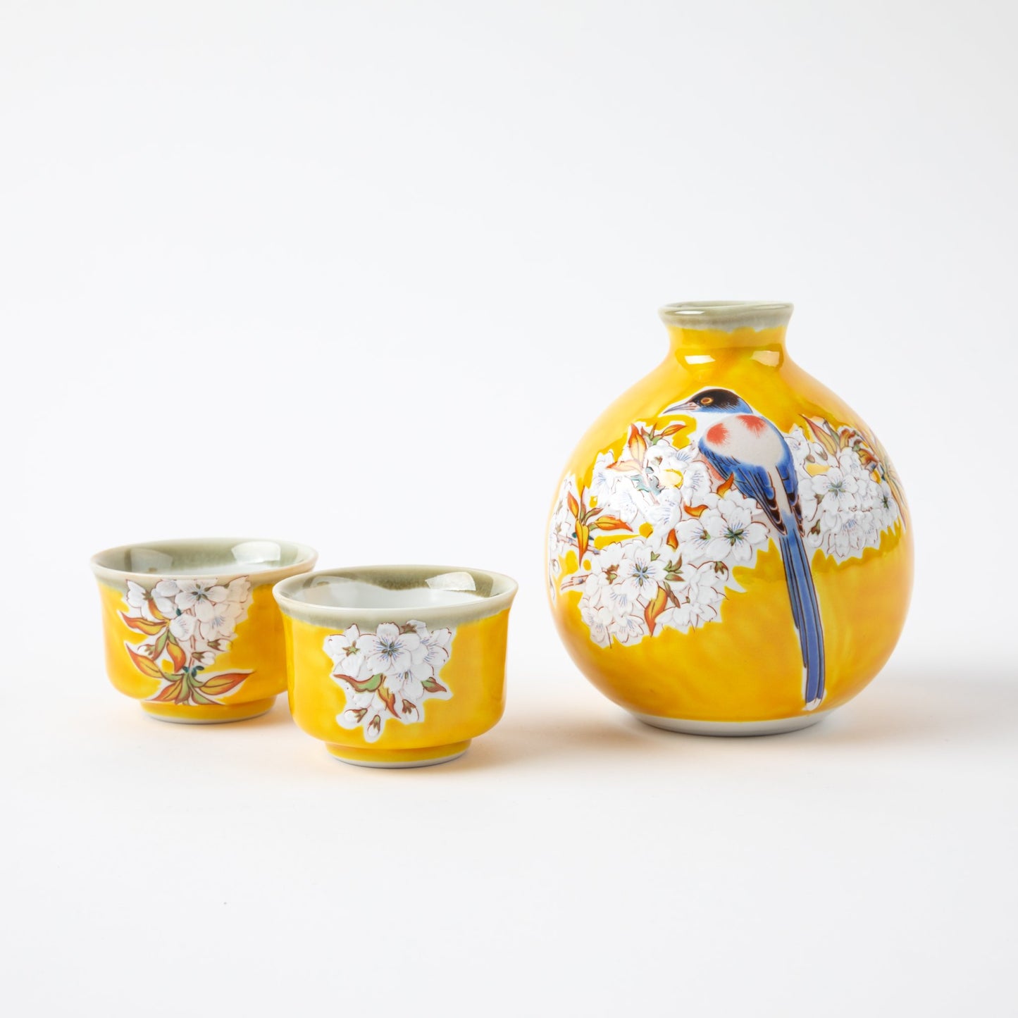 A vibrant yellow sake bottle featuring a detailed blue bird perched among blooming white cherry blossoms, accompanied by two matching yellow sake cups adorned with cherry blossom designs.