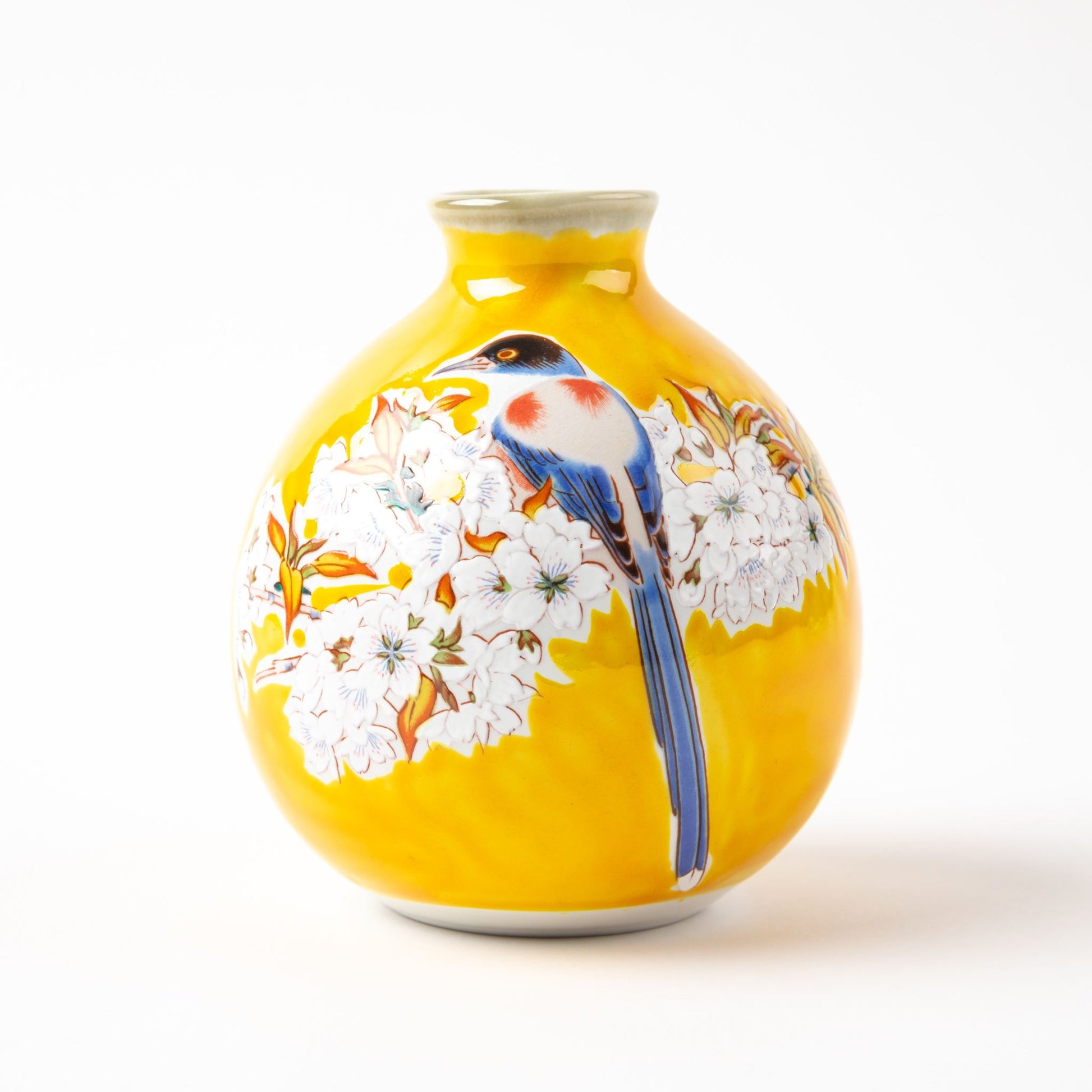 A detailed view of the yellow sake bottle showcasing intricate hand-painted cherry blossoms and a blue bird, highlighting the craftsmanship of the Kutani Yoshidaya design.