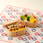 A close-up of a wooden bento box filled with various Japanese dishes, placed on a patterned cloth.