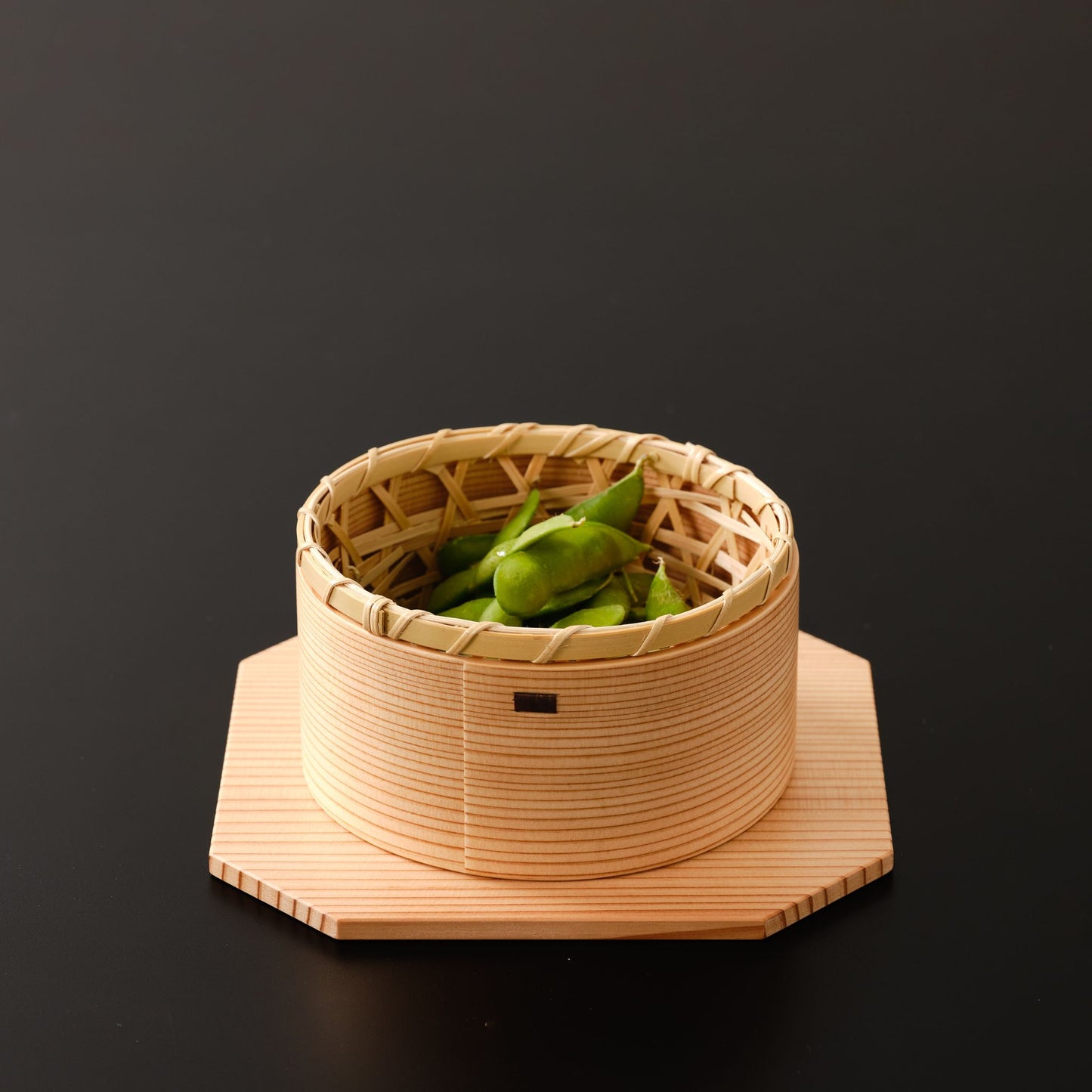 A small bamboo basket with green beans, placed inside a wooden bowl on an octagonal base.