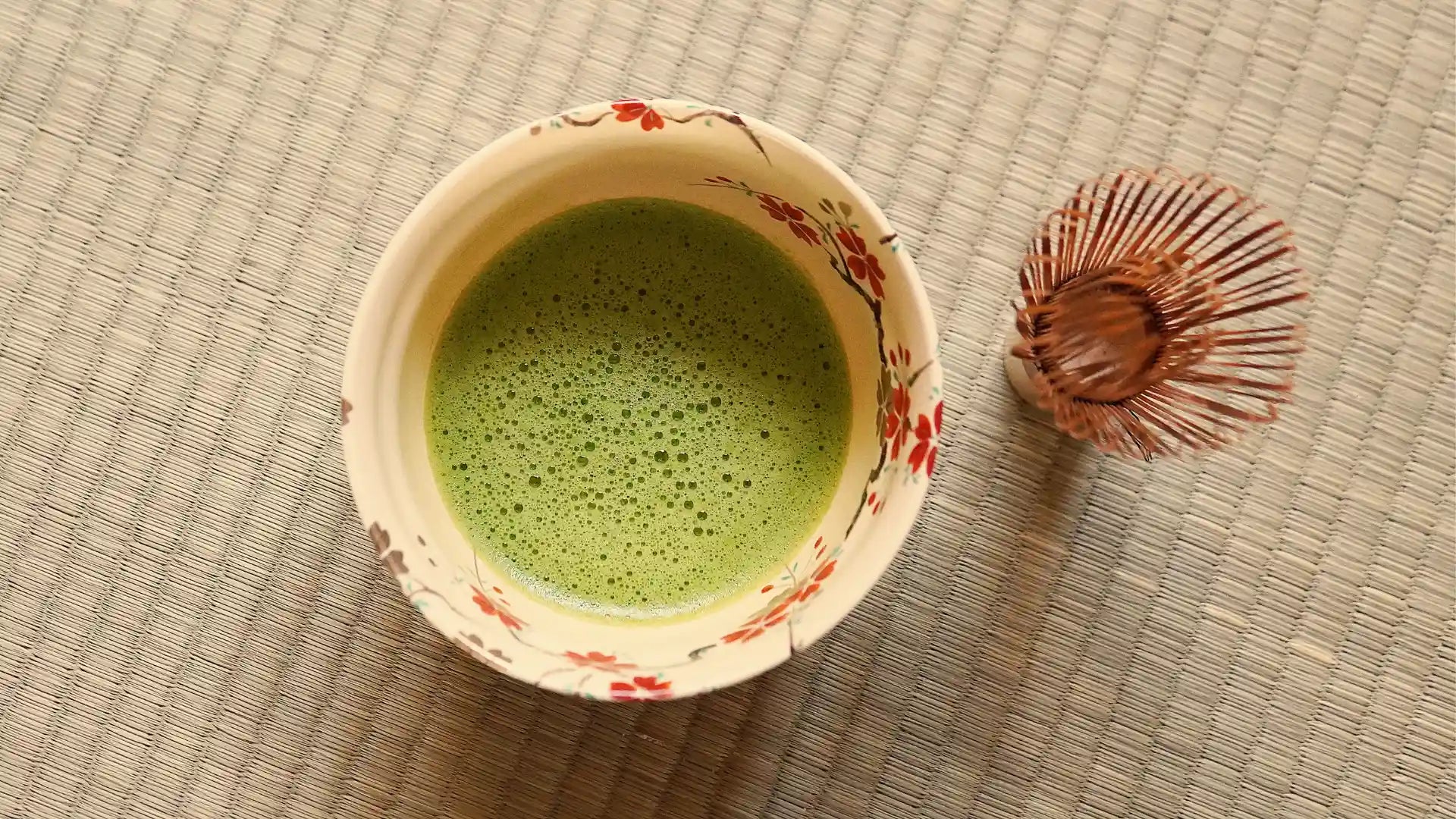 A traditional ceramic bowl with floral designs filled with frothy green matcha tea, accompanied by a bamboo whisk on a tatami mat.