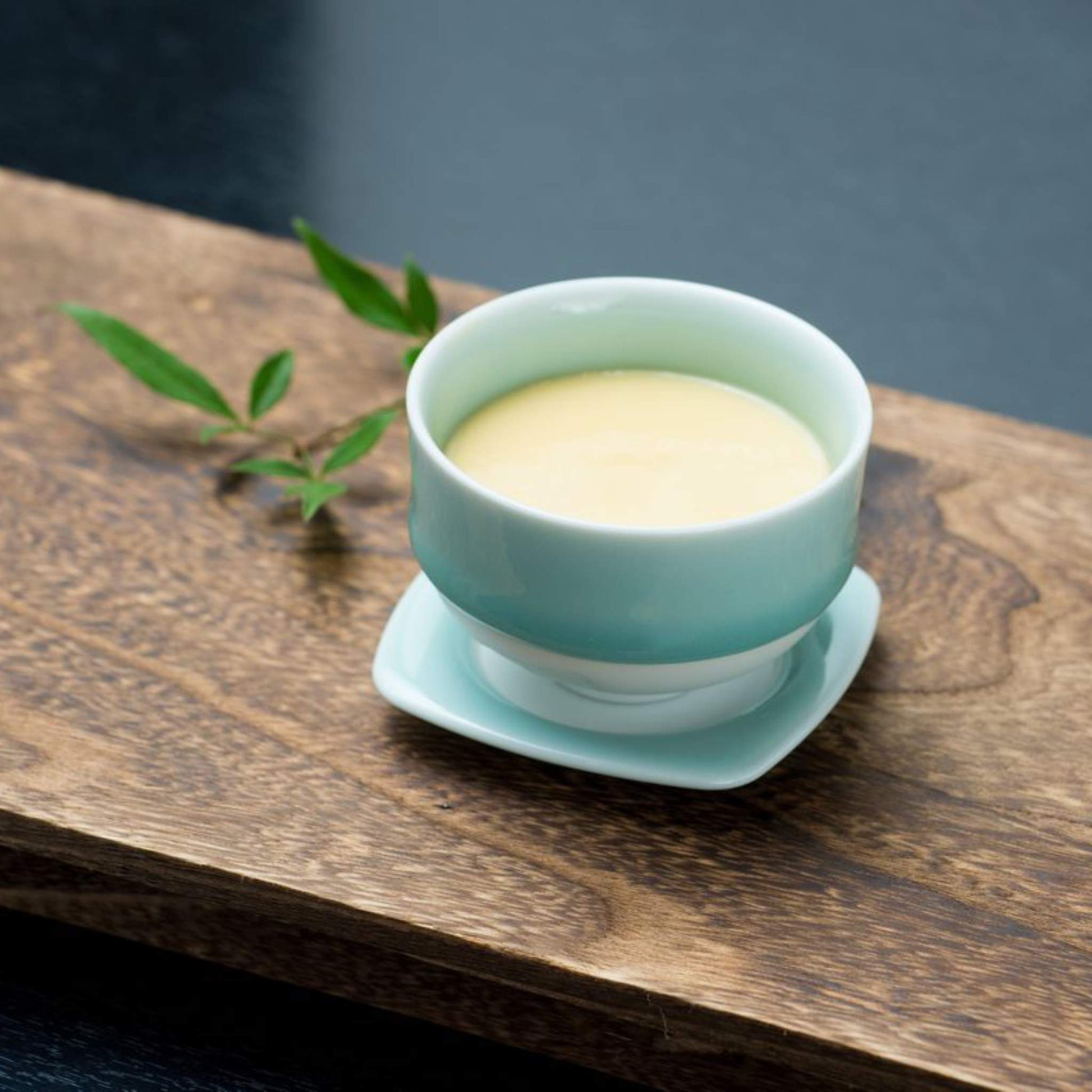 A Nabeshima celadon porcelain tea cup filled with a creamy dessert, placed on a wooden tray with a garnish, highlighting its aesthetic appeal.