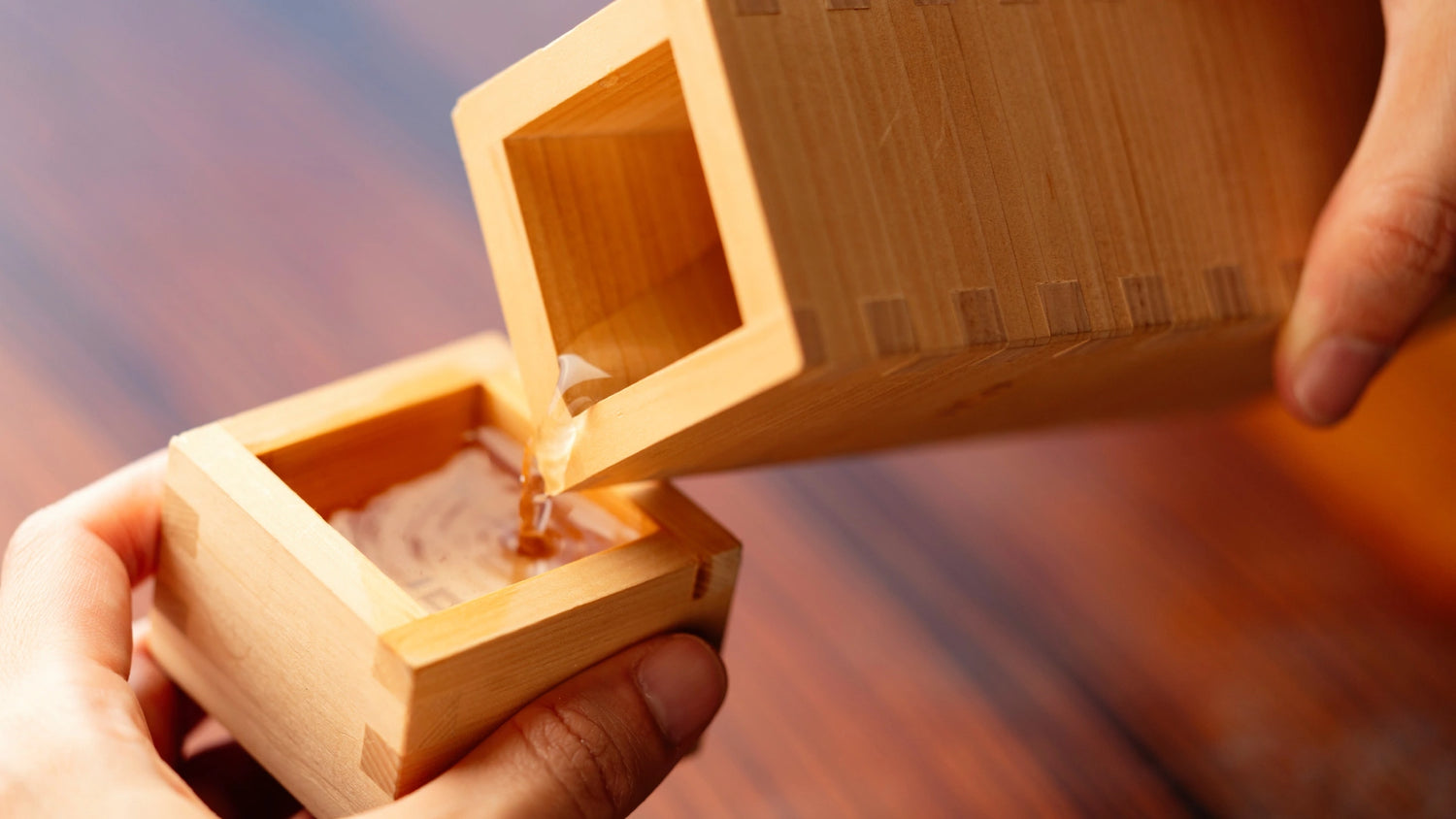 Close-up of sake being poured from a tall wooden tokkuri bottle into a matching square wooden ochoko cup, highlighting the craftsmanship and natural wood grain.