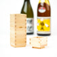 A wooden sake set including an ochoko cup and a tokkuri bottle in front of two blurred sake bottles with colorful labels.