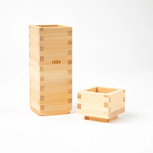 A pair of matching wooden pieces, a square ochoko cup and a tall tokkuri bottle, side by side on a white background.