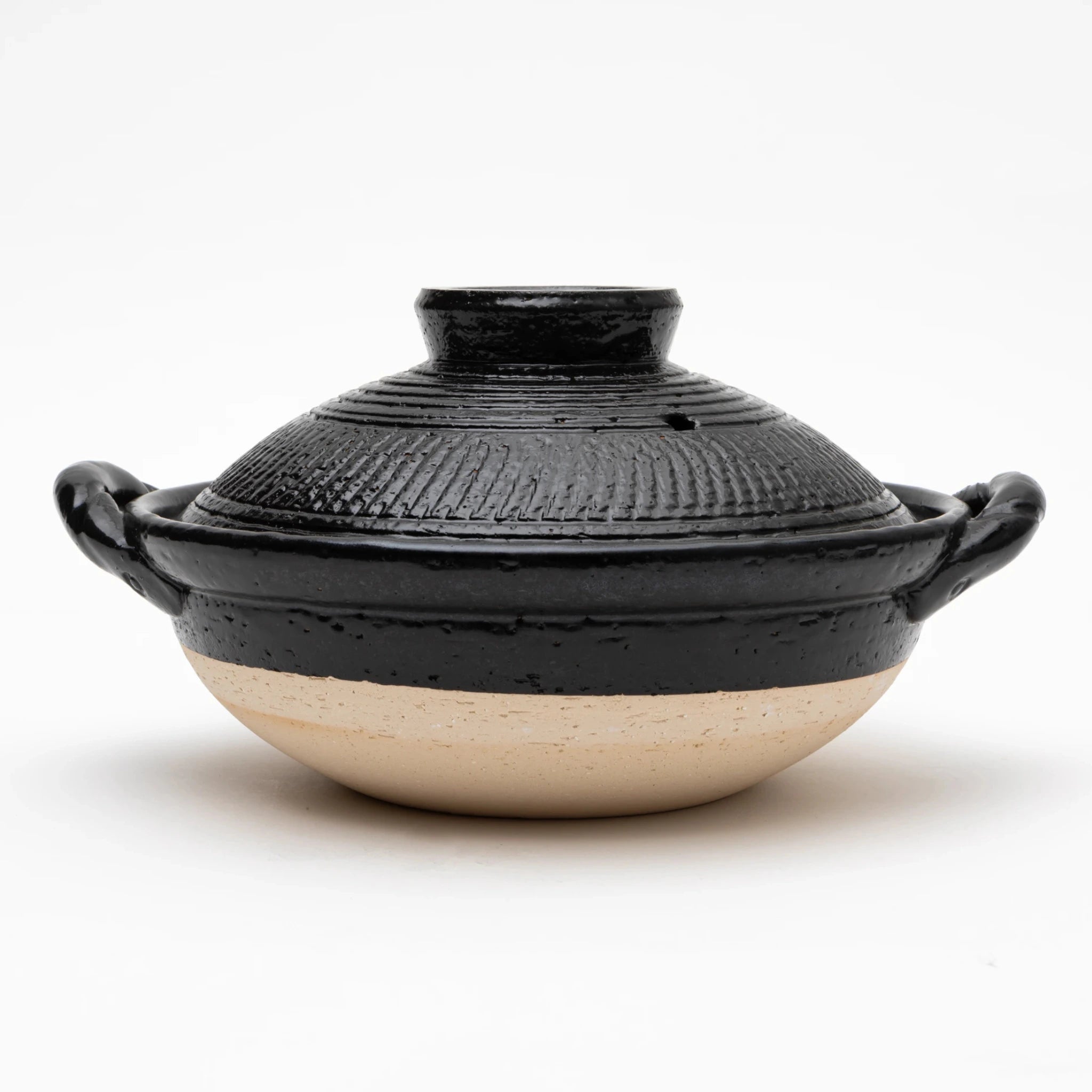 Front view of the Sajitouki Iga ware tobikanna donabe in black, showing a domed lid with fine parallel chattering tool grooves under black glaze, looped side handles, and an unglazed pale clay lower body.