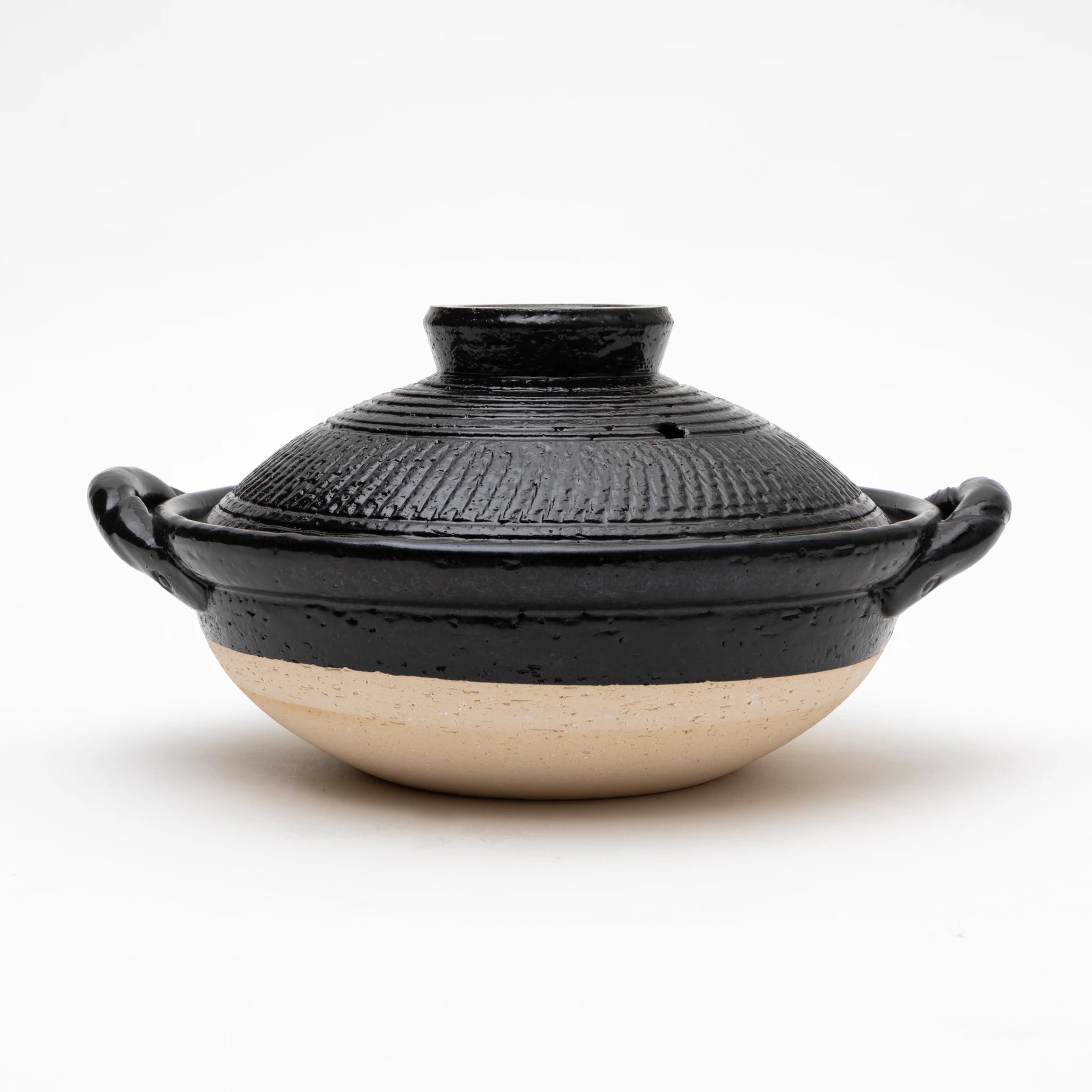 Three-quarter view of the Sajitouki Iga ware tobikanna donabe in black, showing a domed lid with fine parallel chattering tool grooves under black glaze, looped side handles, and an unglazed pale clay lower body.