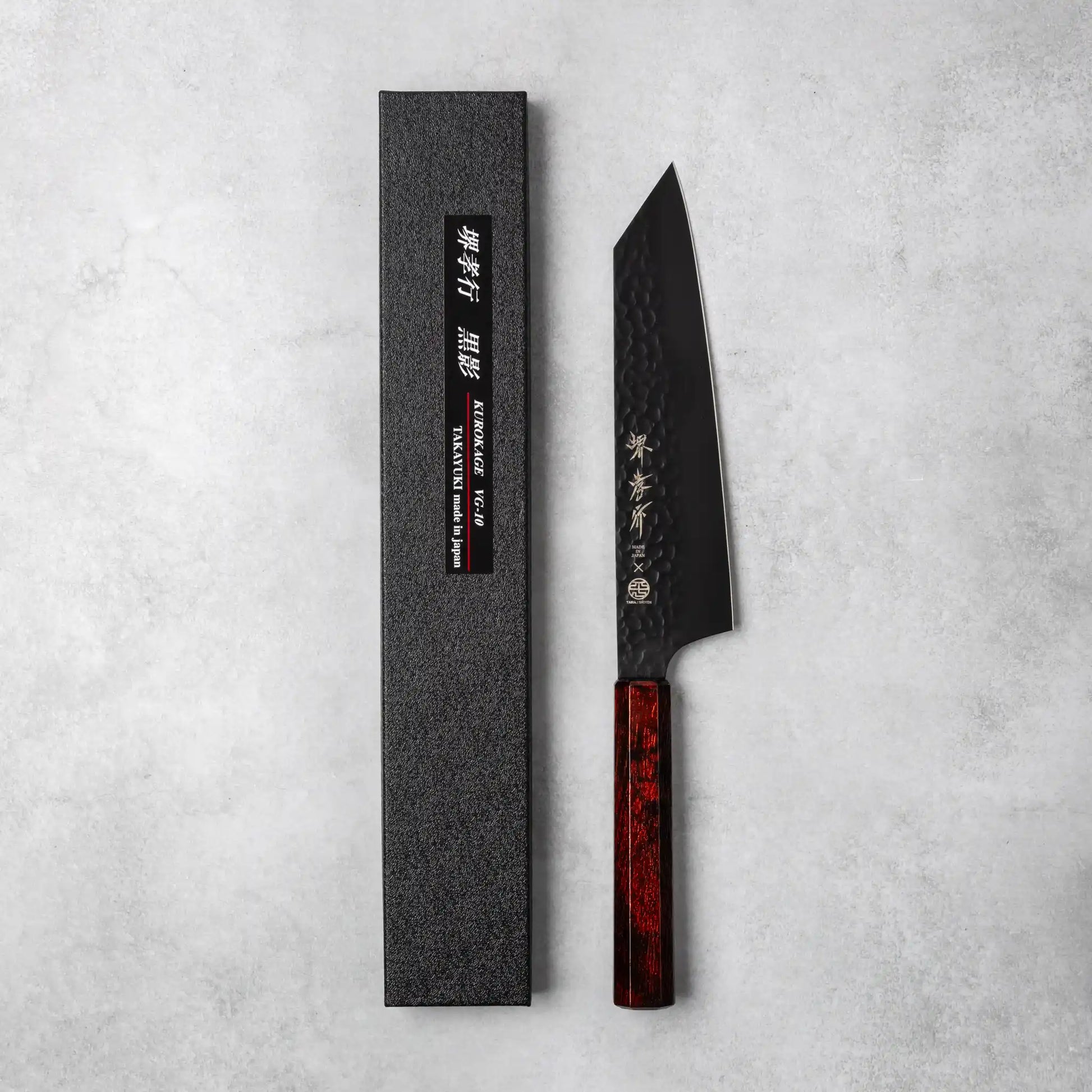 A Sakai Takayuki Kurokage VG10 Kengata chef knife with a hammered black blade and a rich burgundy resin octagonal handle, displayed alongside its black textured packaging on a neutral background.