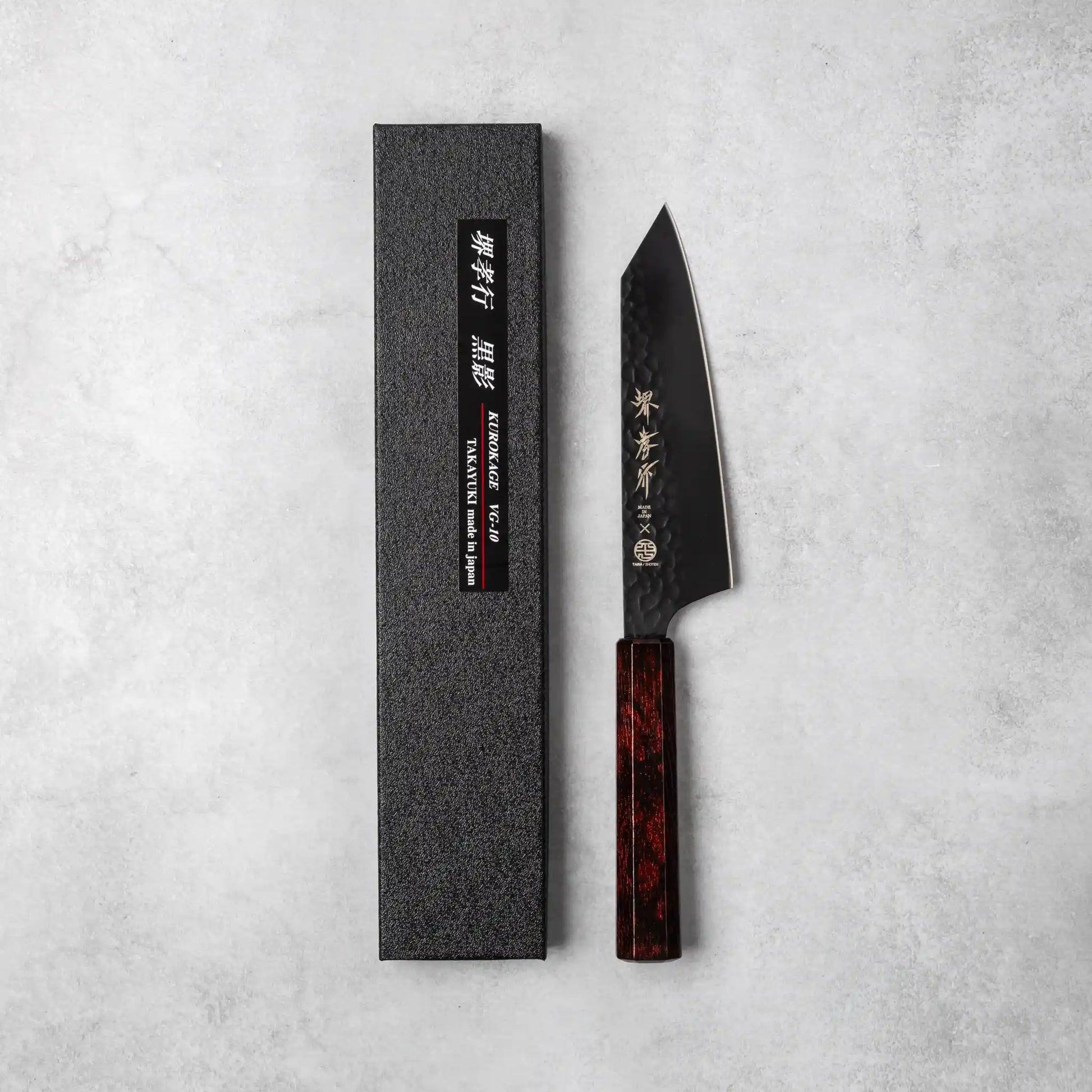 Product box and Sakai Takayuki Kurokage VG10 Kengata Santoku Knife with a hammered black blade and burgundy handle on a textured gray surface.