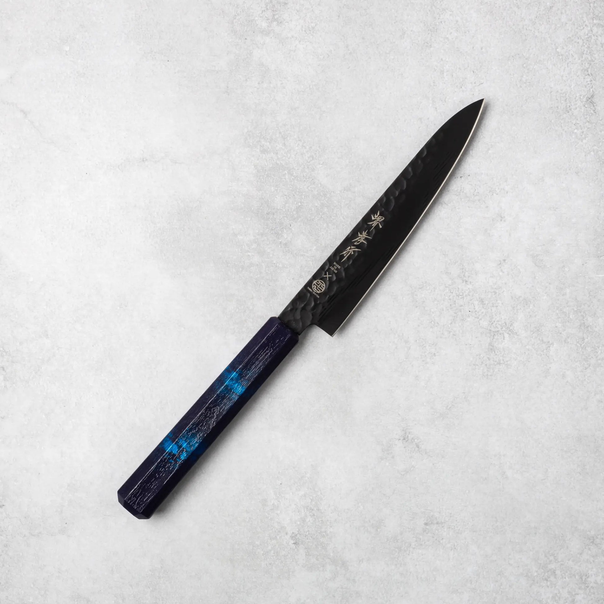 Sakai Takayuki Kurokage VG10 petty knife, showcasing its hammered black blade and smooth blue resin handle, positioned diagonally on a light stone surface.