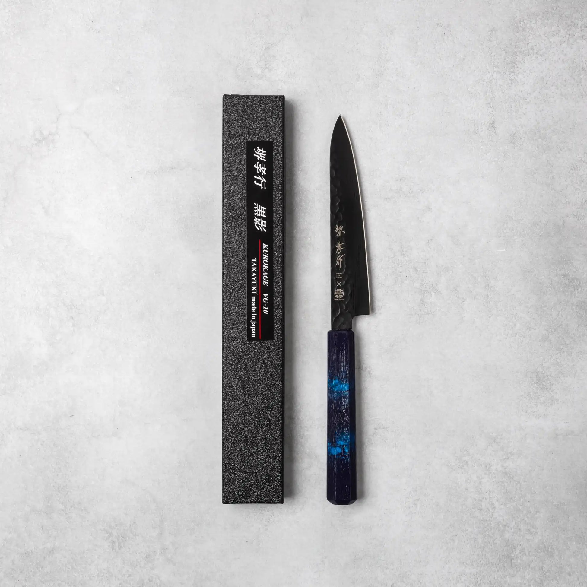 A Sakai Takayuki Kurokage VG10 petty knife with a hammered black blade and an elegant blue resin octagonal handle, alongside its black textured packaging on a neutral background.