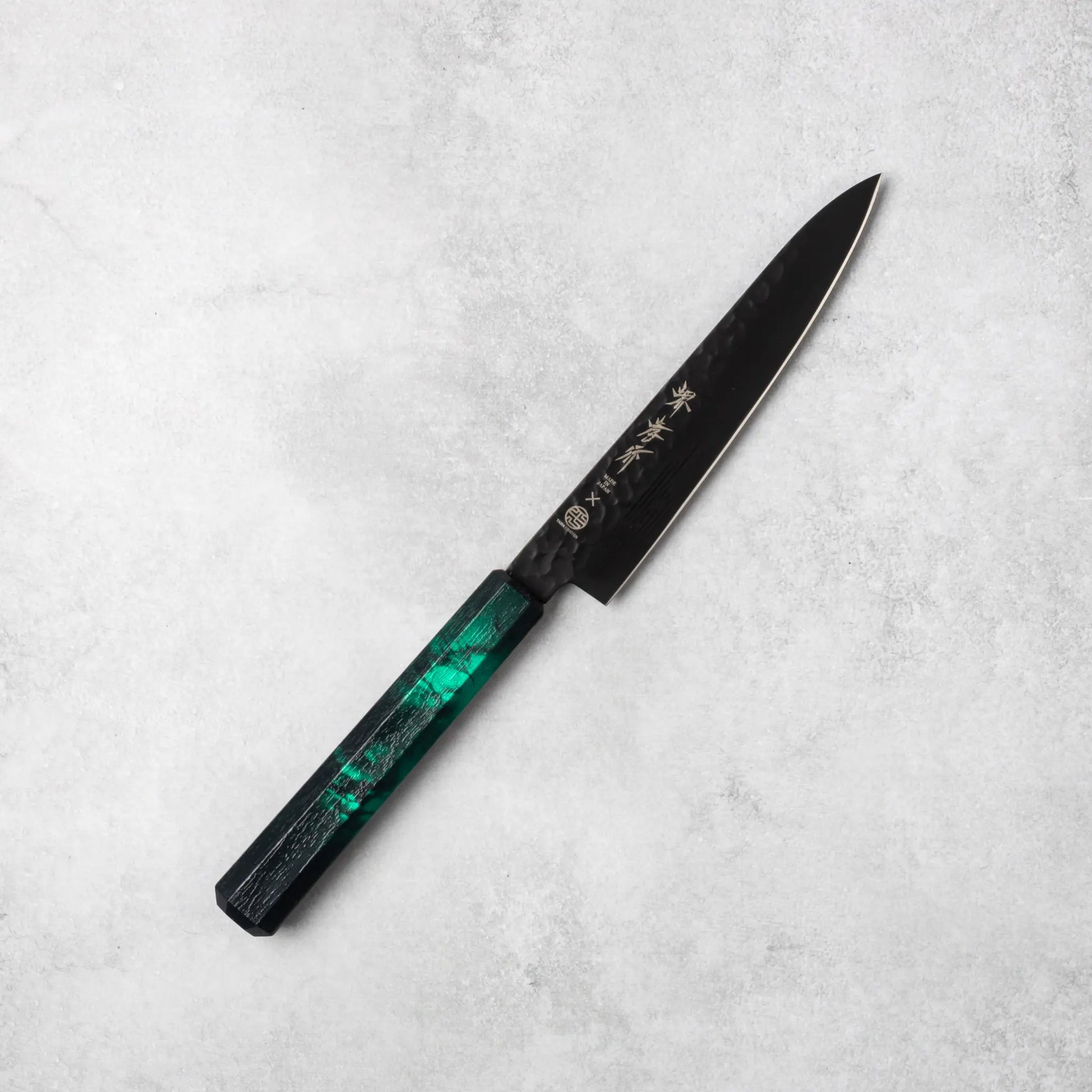 Sakai Takayuki Kurokage VG10 petty knife, featuring a hammered black blade and vibrant green resin handle, positioned diagonally on a light stone surface.