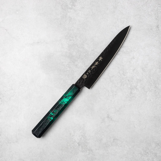 Sakai Takayuki Kurokage VG10 petty knife, featuring a hammered black blade and vibrant green resin handle, positioned diagonally on a light stone surface.