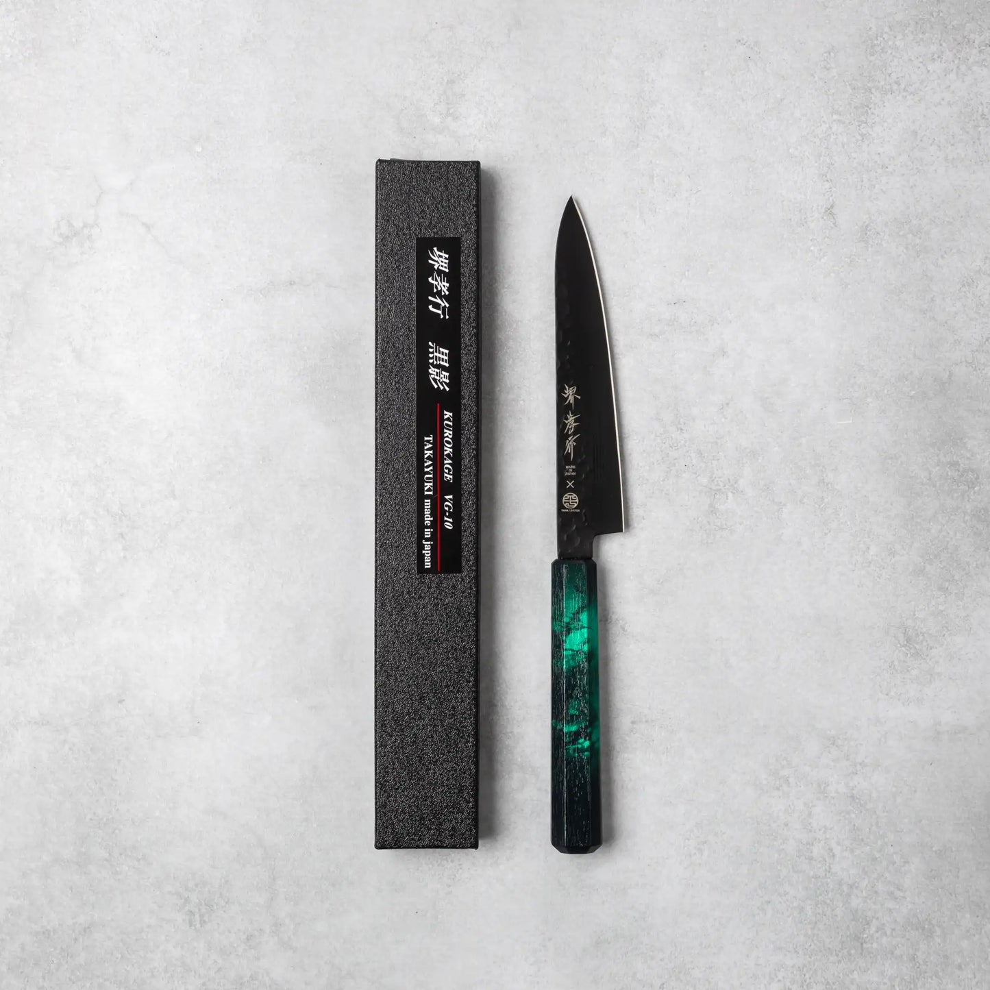 A Sakai Takayuki Kurokage VG10 petty knife with a hammered black blade and a striking green resin octagonal handle, displayed with its black textured packaging on a neutral background.