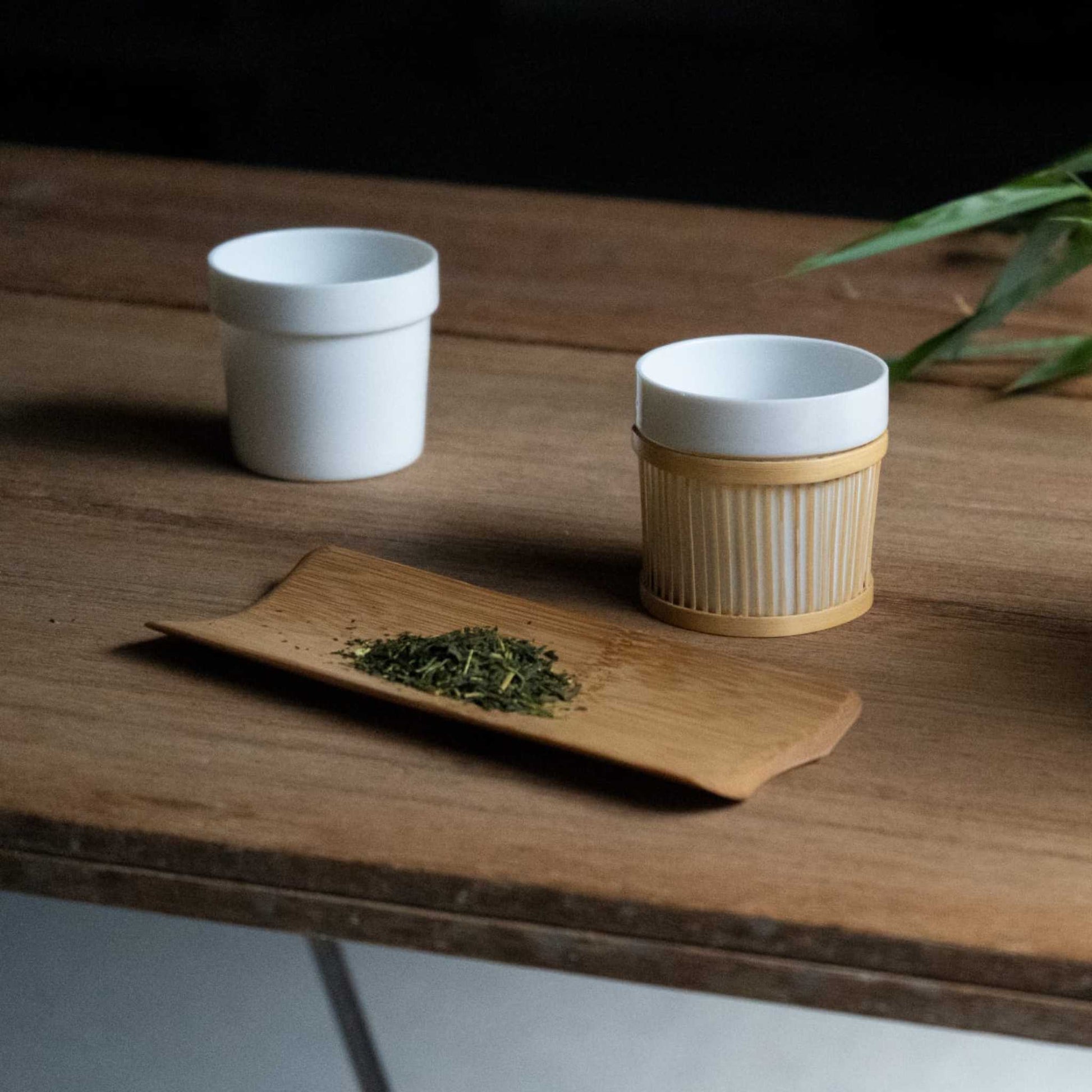 Two Sen Bamboo Sleeve Cups, one with a bamboo sleeve and one without, placed on a wooden table alongside a bamboo tea scoop filled with green tea leaves.