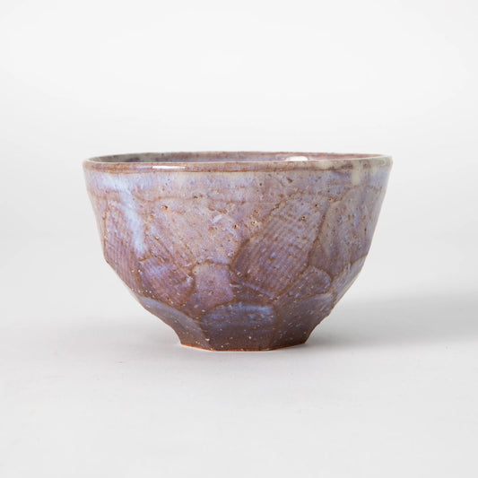 A Hagi ware matcha bowl by Senryuzan with broad Misogi carved exterior facets beneath a white slip and translucent glaze in dusty lavender and muted rose tones, with warm iron clay visible through the glaze at the ridges, photographed against a white background.