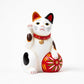 Front-facing Maneki Neko (beckoning cat) in a traditional ceramic design, showcasing white fur with multicolored spots and a red collar with a bell. The cat raises its left paw and holds a decorative red ball with intricate patterns, symbolizing luck and prosperity.