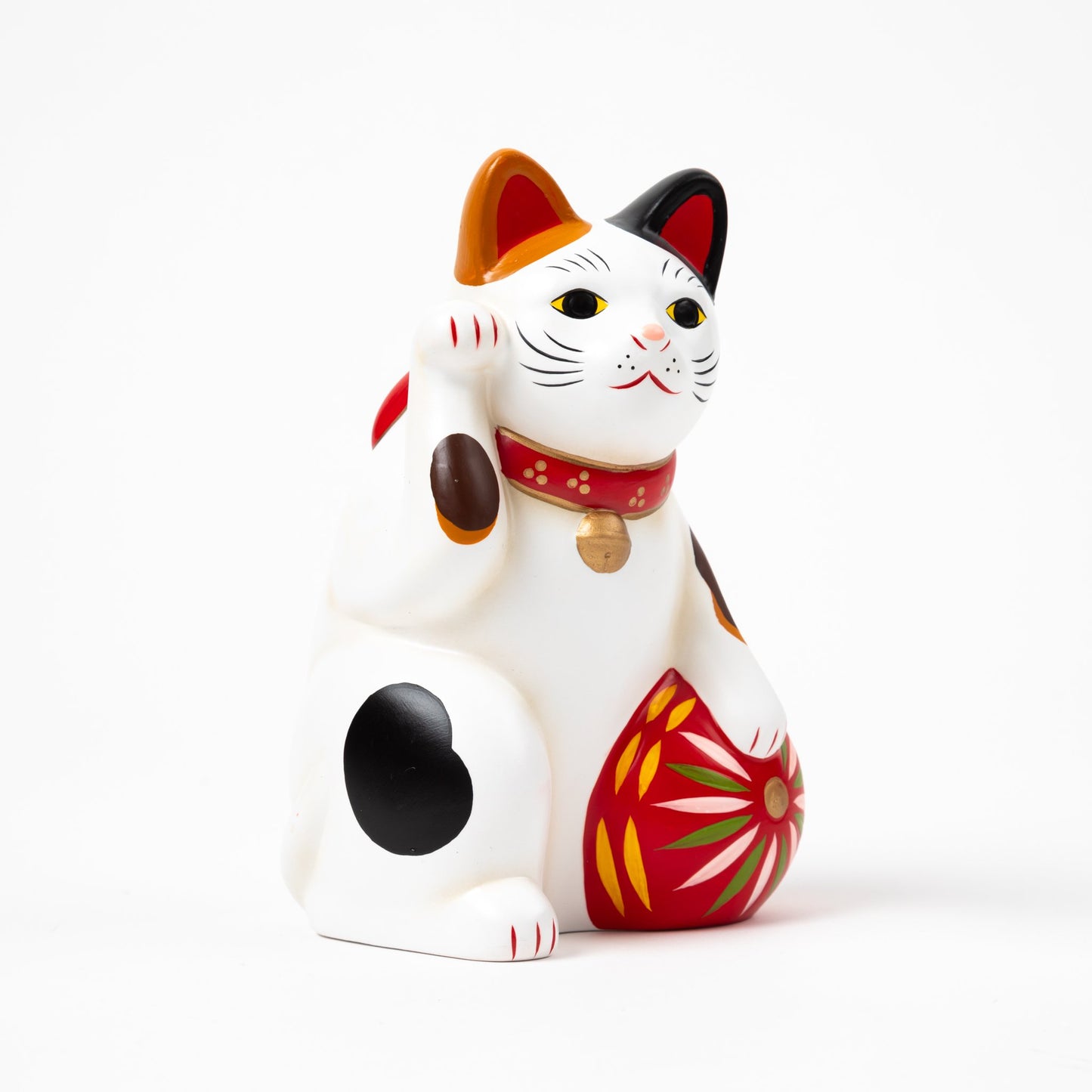 Righ-side profile of a classic ceramic Maneki Neko (beckoning cat) raising its left paw, with white fur and black, orange, and brown spots. The cat wears a red collar with a bell and holds a vibrant red ball decorated with colorful patterns.