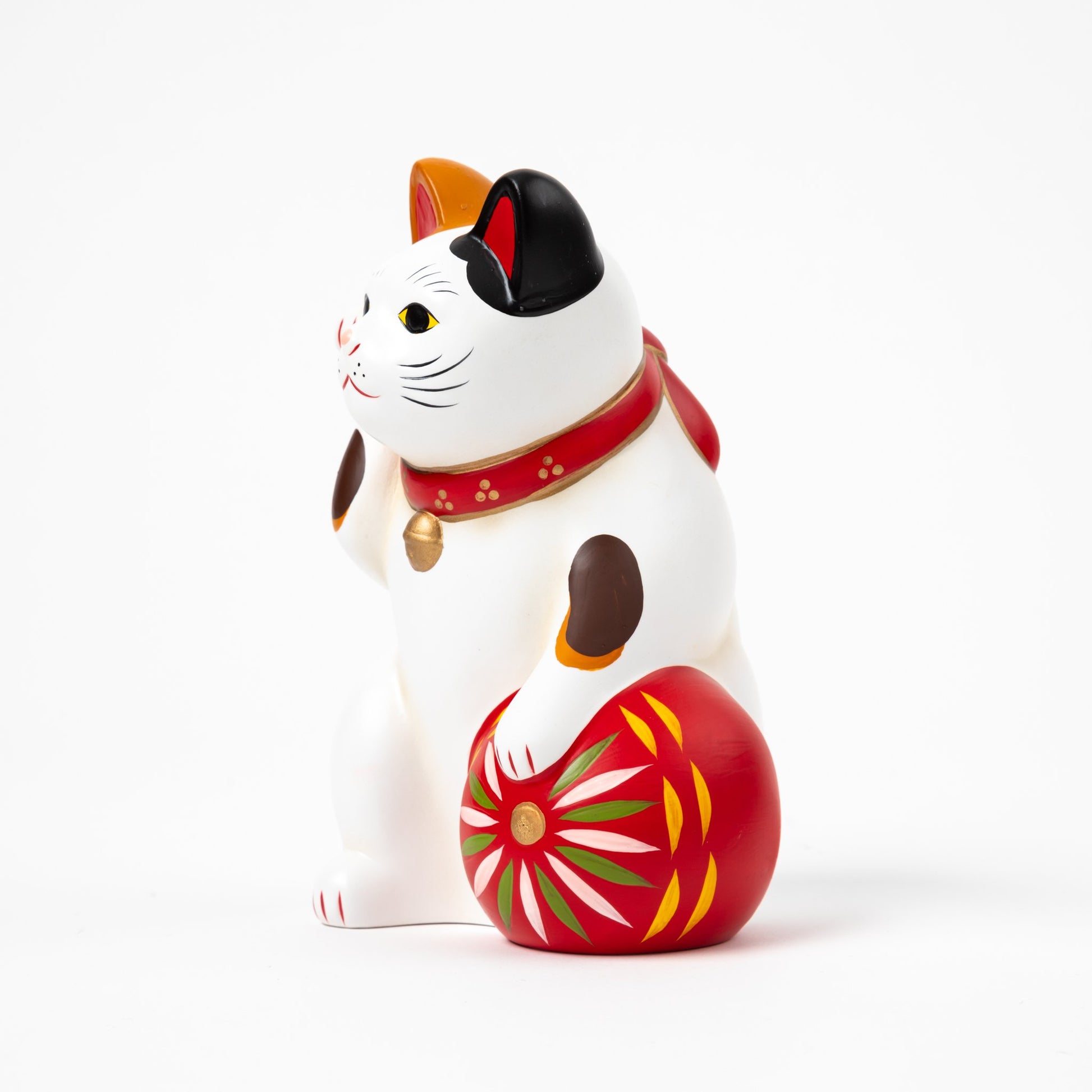 Side profile of a traditional ceramic Maneki Neko (beckoning cat) with white fur and multicolored spots, wearing a red collar and bell. The cat holds a decorative red ball featuring colorful patterns, symbolizing good fortune.