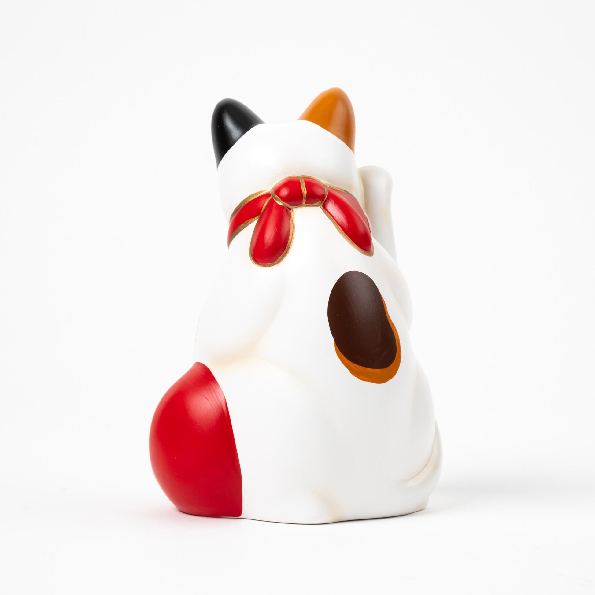 Back view of a ceramic Maneki Neko (beckoning cat) in a traditional design, featuring white fur with black, orange, and brown spots. The cat is adorned with a red collar and bell, holding a vibrant red ball at its side.