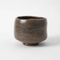 A hand built black raku matcha bowl by Shoraku Kiln in the Chojiro Kamuro style, with a cylindrical tapered form, irregular rim, and a deep iron dark glaze showing horizontal striations and warm brown tones, photographed against a white background.