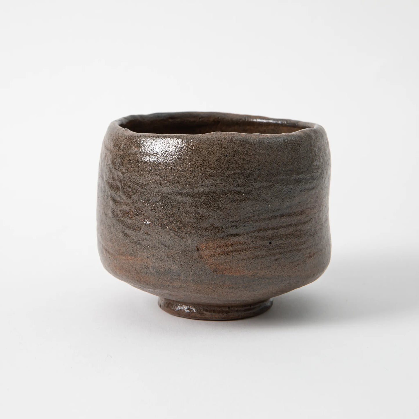 A hand built black raku matcha bowl by Shoraku Kiln in the Chojiro Kamuro style, with a cylindrical tapered form, irregular rim, and a deep iron dark glaze showing horizontal striations and warm brown tones, photographed against a white background.