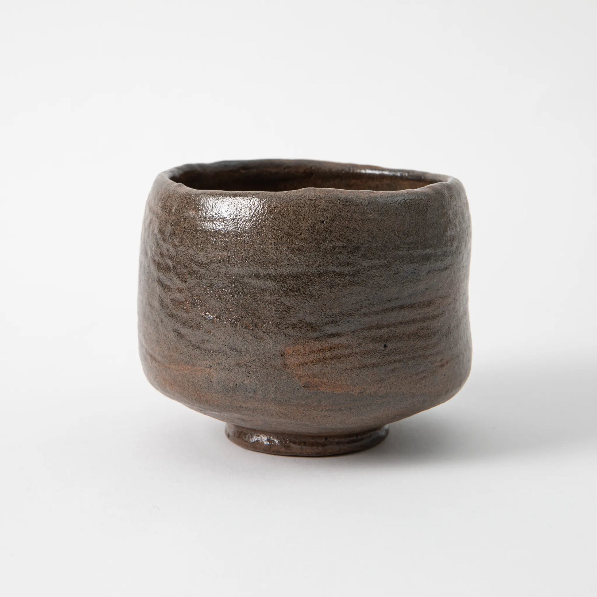 A hand built black raku matcha bowl by Shoraku Kiln in the Chojiro Kamuro style, with a cylindrical tapered form, irregular rim, and a deep iron dark glaze showing horizontal striations and warm brown tones, photographed against a white background.