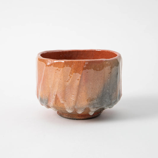 A hand built red raku matcha bowl by Shoraku Kiln with vertical ridges, a wide irregular form, and a glaze in warm coral, pale blush, and soft grey tones with crystalline drip edges at the lower body, photographed against a white background.Sonnet 4.6