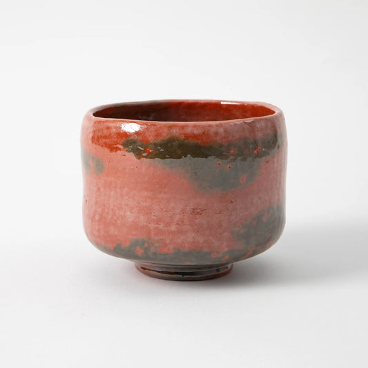 A hand built red raku matcha bowl by Shoraku Kiln in the Sonyu Hanaikada style, with a low settled form, deep terracotta red glaze shifting to darker iron passages, and a small grounded foot, photographed against a white background.