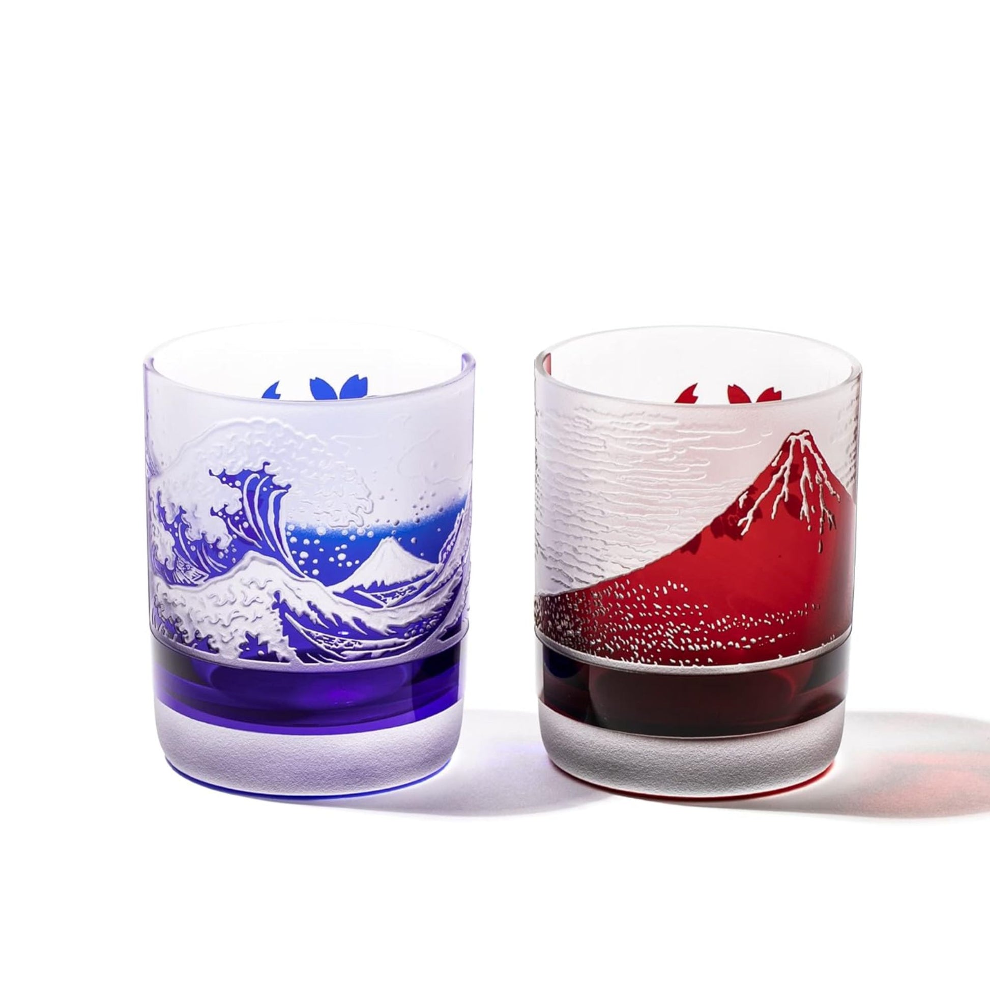 Two intricately designed whiskey glasses side by side. One glass features a blue depiction of Mount Fuji and a crashing wave, while the other displays a red Mount Fuji with a textured design. Both glasses incorporate sakura (cherry blossom) details, showcasing fine Japanese craftsmanship.