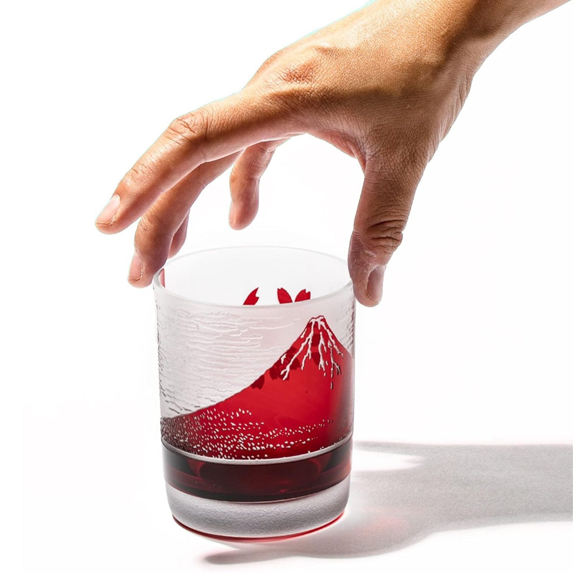 A hand reaching out to hold the red whiskey glass adorned with Mount Fuji and sakura designs, showing the scale of the glass.