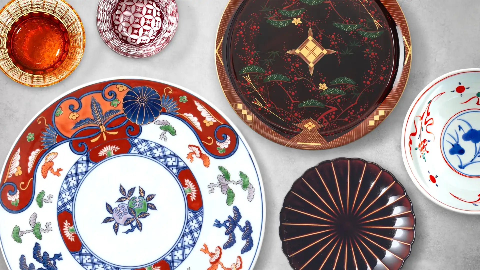 A collection of traditional Japanese tableware, including vibrant porcelain plates, lacquerware trays, and glass bowls, arranged on a minimalist gray background.