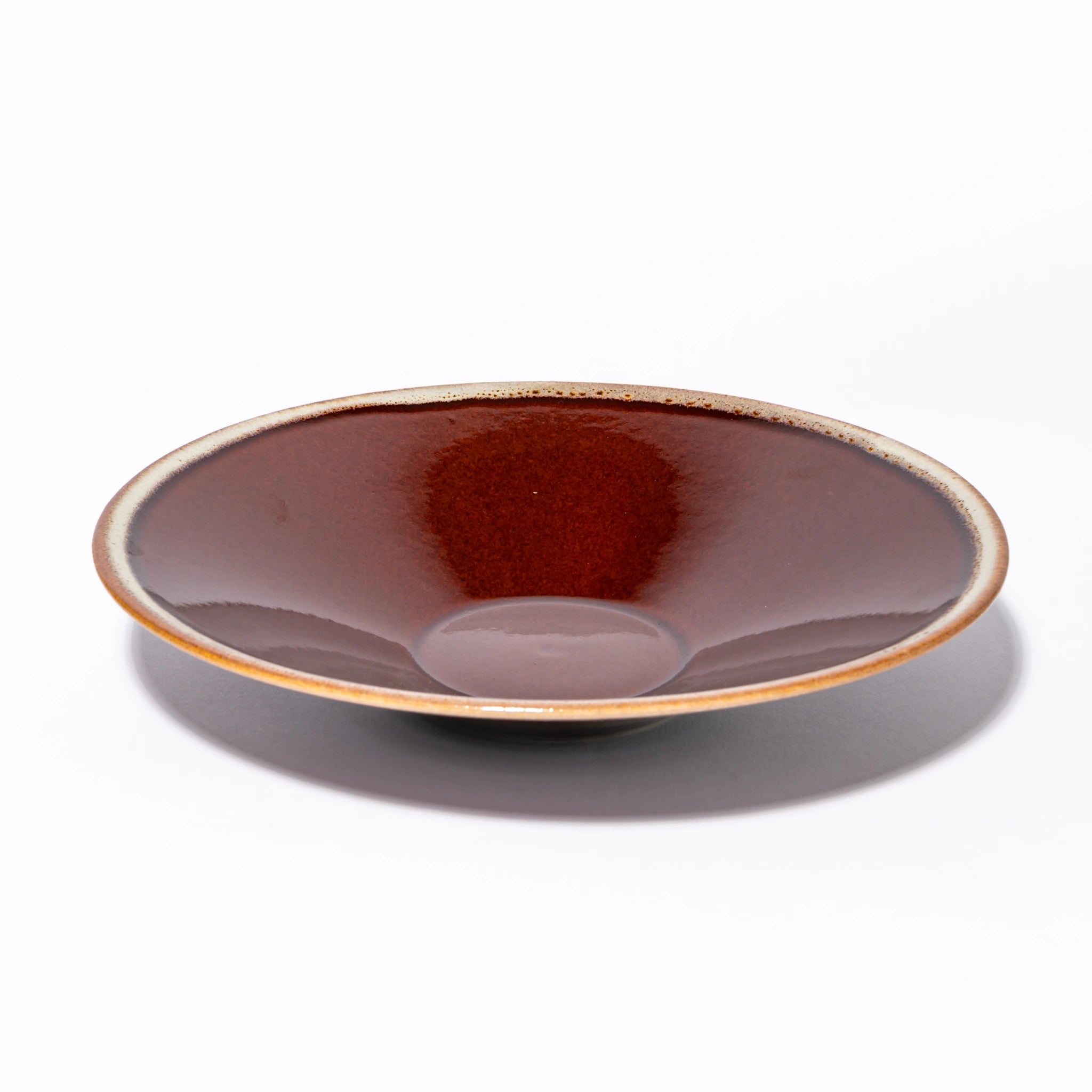 Three-quarter view of the Tsukamoto Mashiko ware SABI flat bowl large in amber glaze, showing a wide shallow form with deep iron red glaze at the center transitioning to pale grey-cream at the rim with amber iron spotting at the edge.