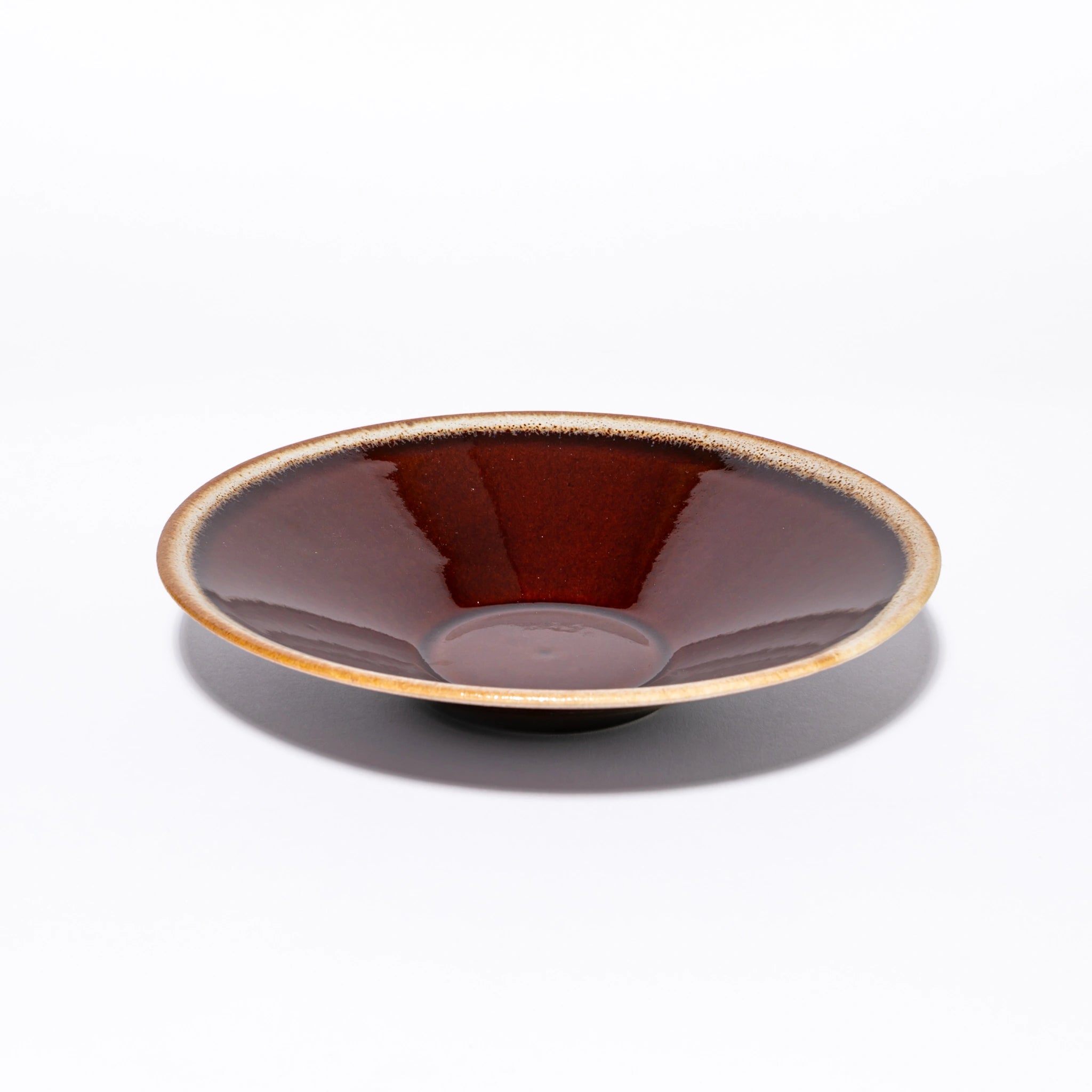Three-quarter view of the Tsukamoto Mashiko ware SABI flat bowl medium in amber glaze, showing a wide shallow form with deep iron red glaze at the center transitioning to pale grey-cream at the rim with amber iron spotting at the edge.