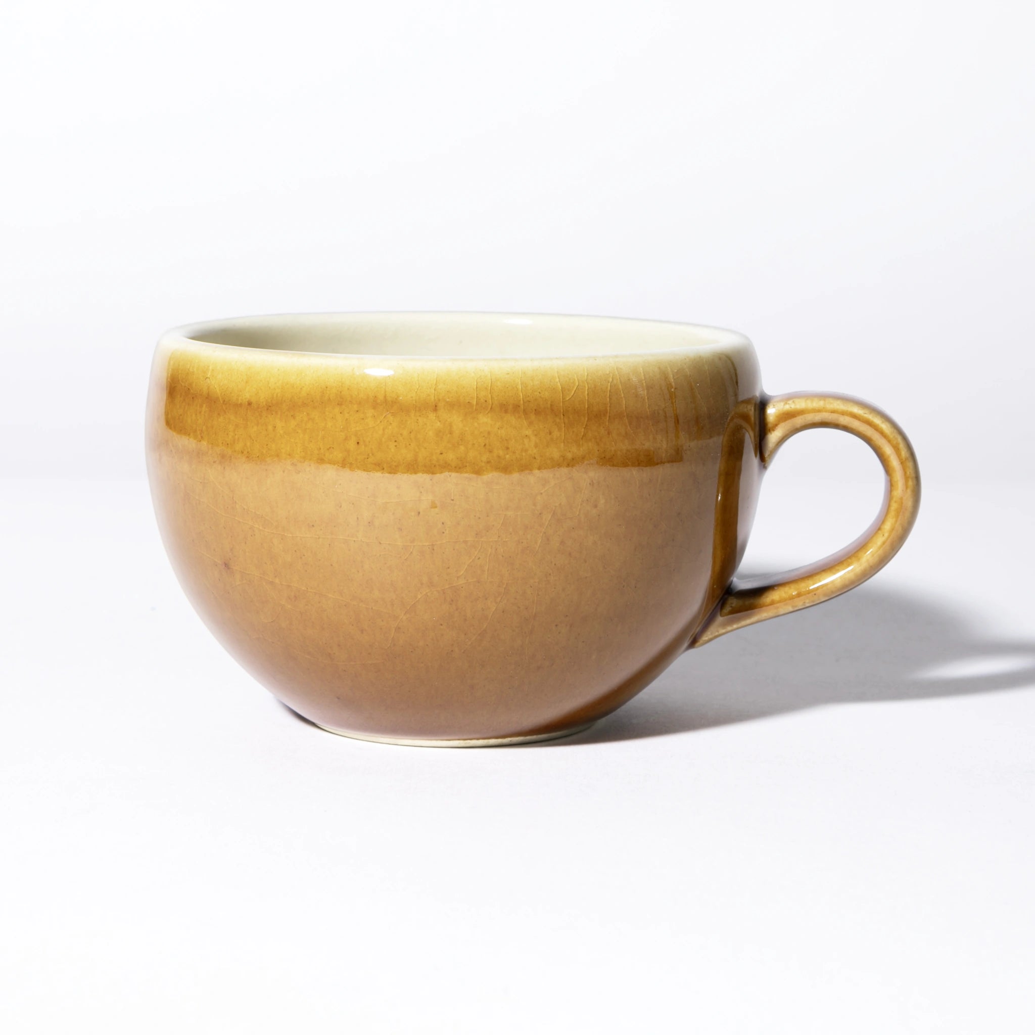 Side view of the Tsukamoto Mashiko ware soup cup in amber glaze, showing a wide rounded form with a loop handle, glaze transitioning from pale cream at the rim through golden amber to rosy brown at the base, with fine crazing across the surface.