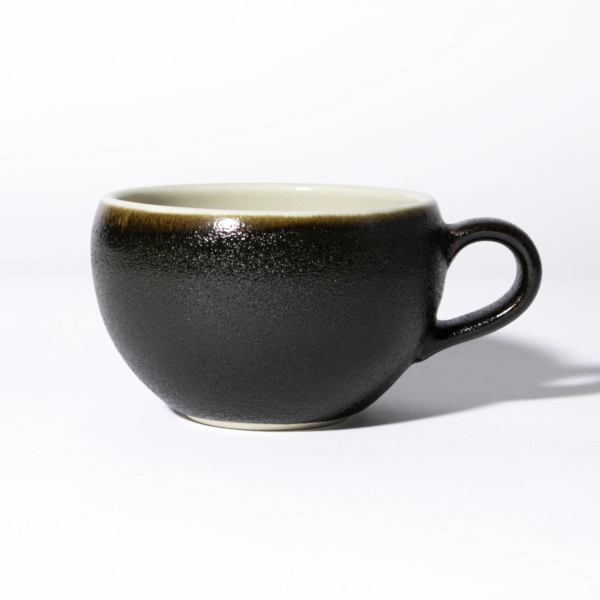 Side view of the Tsukamoto Mashiko ware soup cup in black glaze, showing a wide rounded form with a loop handle, heavily textured granular black glaze across the body transitioning to warm amber at the rim, and a pale cream interior.