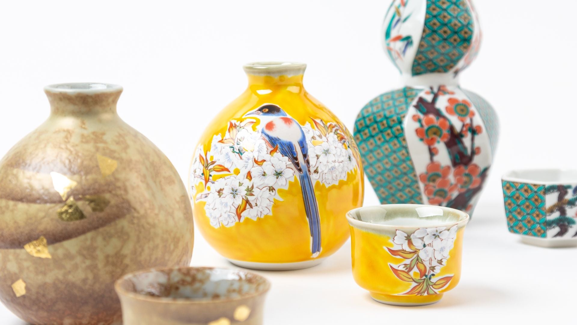 Close-up of a curated display featuring a Kutani sake bottle with a blue bird perched among cherry blossoms on a vibrant yellow background, surrounded by various sake cups and bottles, including a gold-accented sake bottle and a traditional patterned Kutani sake set in green, white, and red hues.