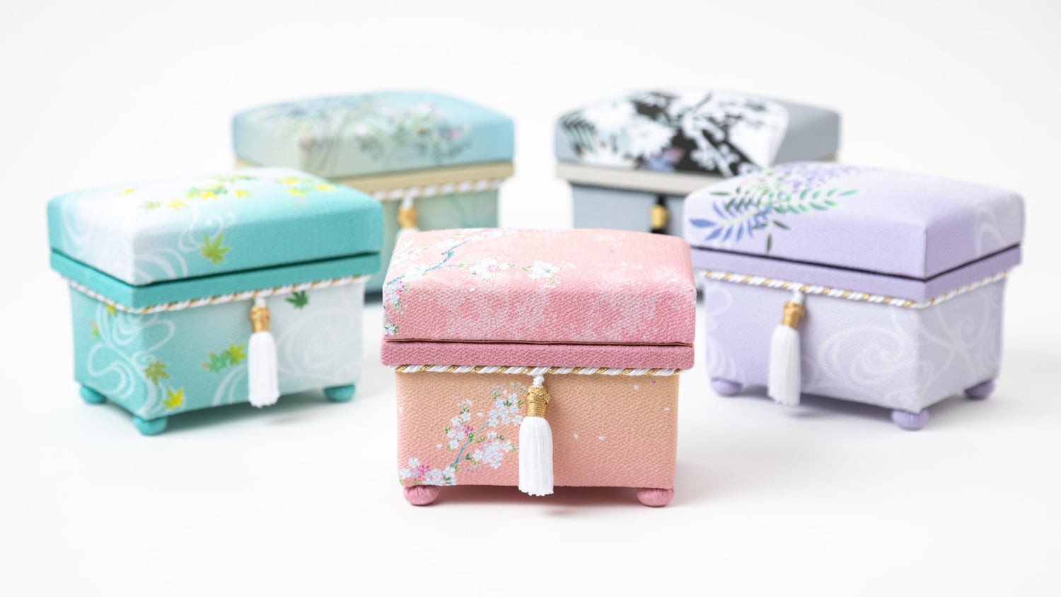 Close-up of hisatsune YUZEN fabric-covered boxes in different pastel shades and floral designs, highlighting the detailed patterns and white tassel embellishments.